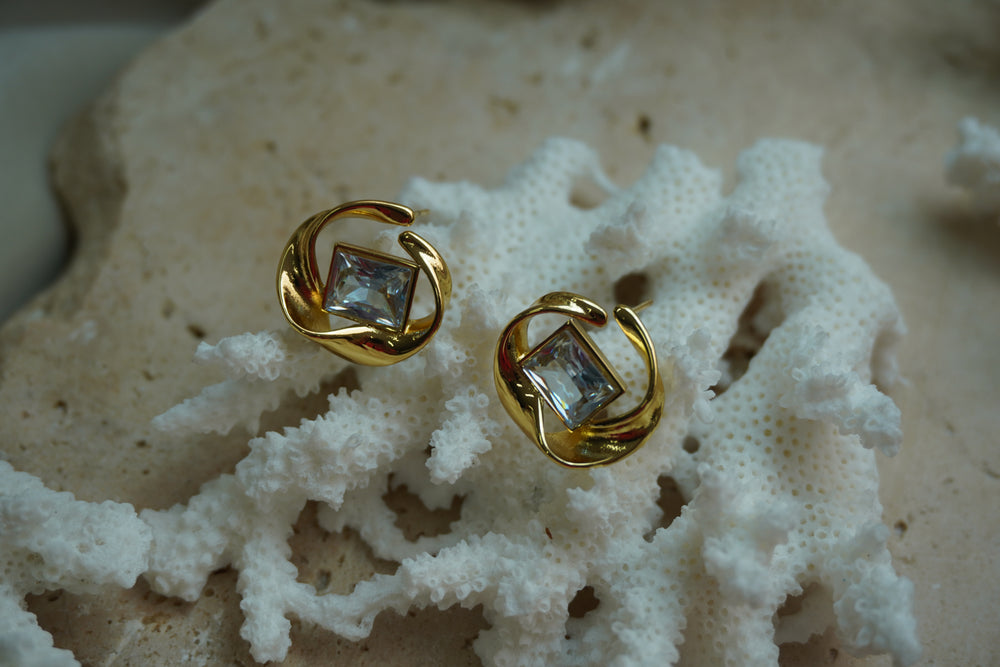 Alternate view: gold-tone stud earrings with clear square centres framed by flowing open circular curves.