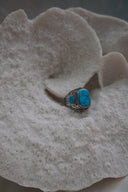 Alternate view: silver-tone ring with a rounded rectangular blue centre, dark markings, circular blue side accents and patterned borders.