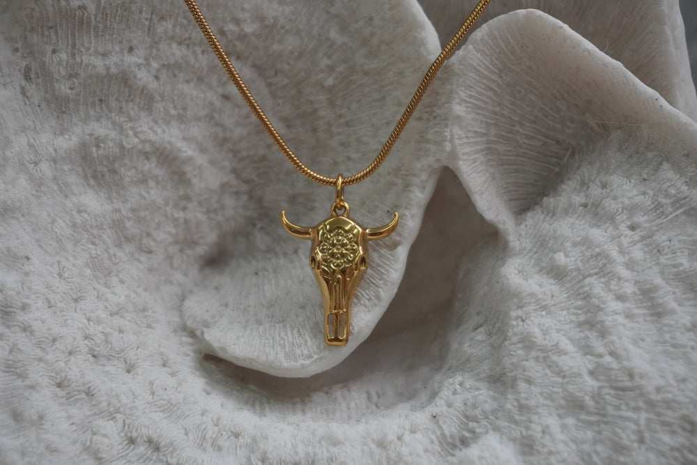 Gold-tone necklace with an elongated skull pendant, floral forehead pattern and outward-curving horns.