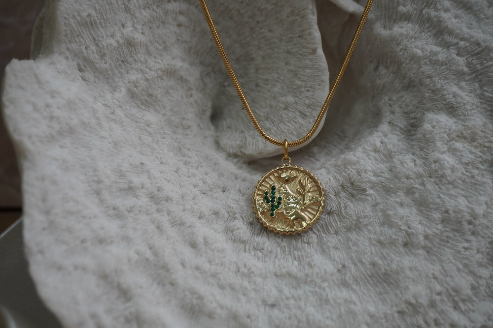 Gold-tone disc necklace with a desert landscape, cactus motifs, green accents and a scalloped pendant edge.