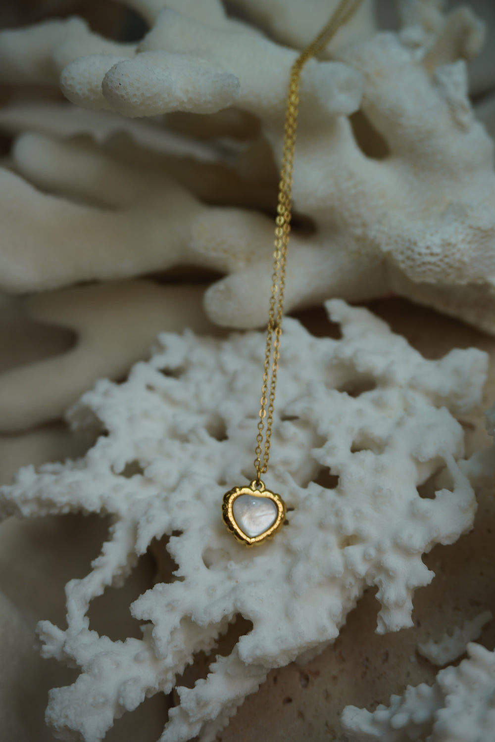 Gold-tone fine chain necklace with a pale iridescent heart centre and a textured, scalloped-looking border.