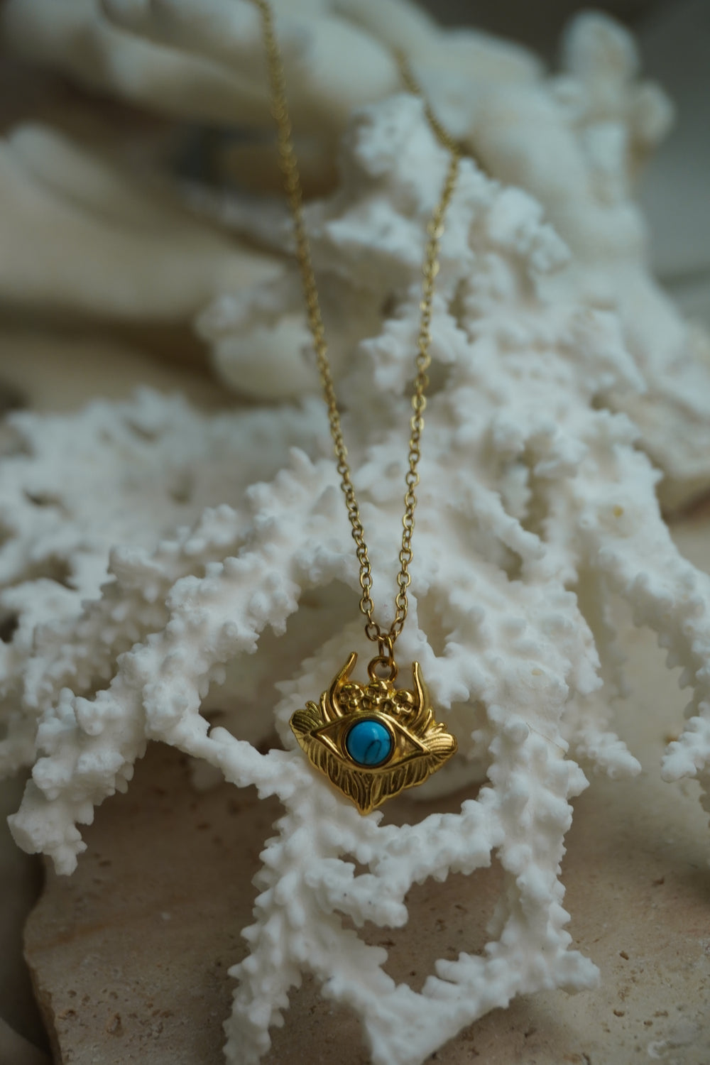 Gold-tone chain necklace with an eye-shaped pendant, bright blue centre, ridged lower border and small skull motifs above.