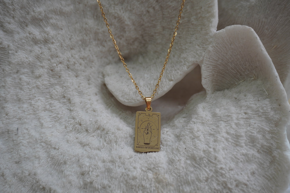 Gold-tone necklace with a rectangular Wheel of Fortune pendant showing crossed fingers and X lettering.
