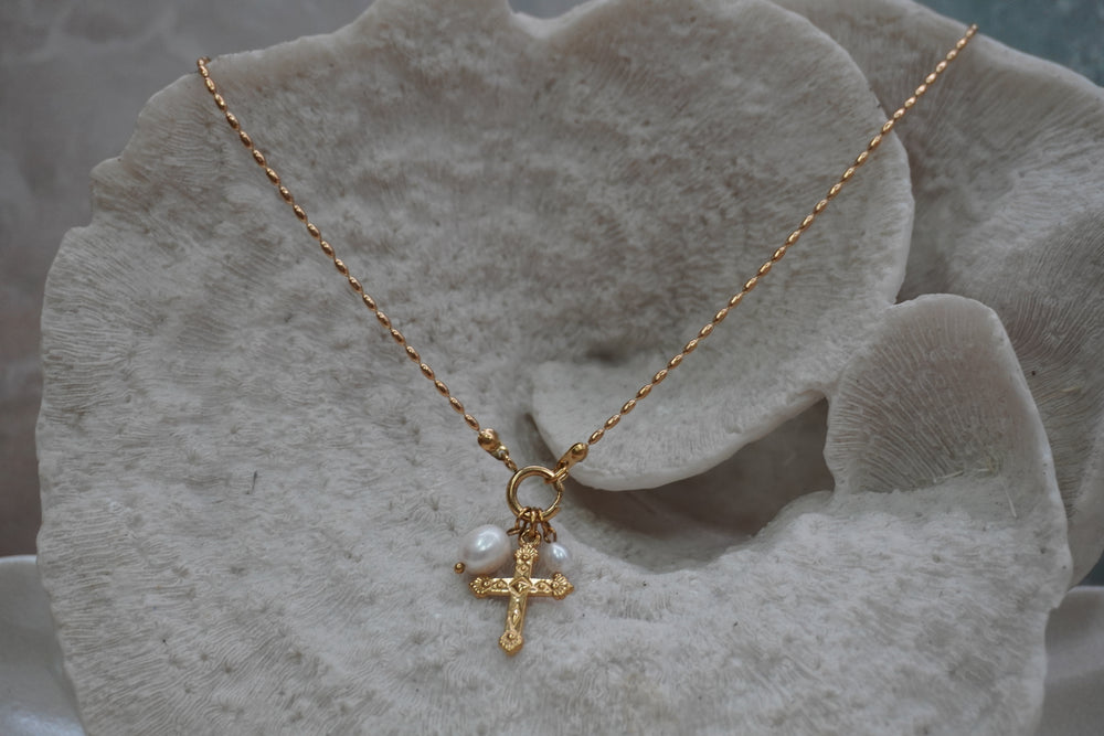 Gold-tone necklace with a patterned cross charm and white oval beads suspended from a front ring.